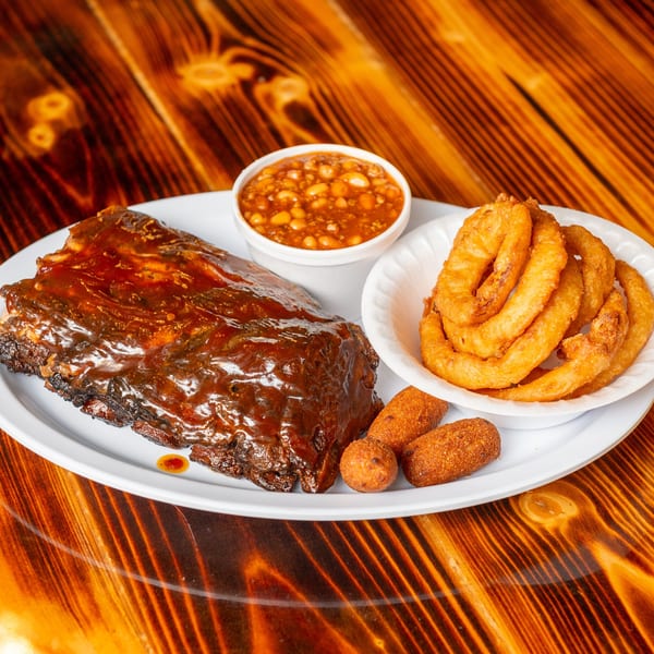 A plate of BBQ ribs, baked beans, onion rings, and hush puppies on a wooden table.
