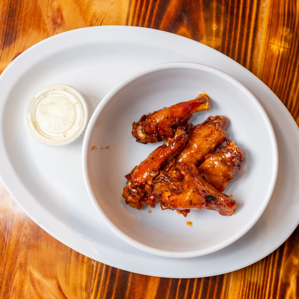 A white bowl of glazed chicken wings and a small cup of dipping sauce on a wooden table.